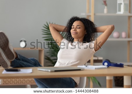 Happy calm african girl student relaxing holding hands behind finished study work breathing fresh air sit at home office desk feel stress relief stretching doing exercise dreaming enjoy peace of mind