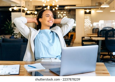 Happy calm brazilian or hispanic female employee resting during work time, relaxed sits at her workplace, puts hands behind head, feels satisfied by project done, looking in distance, smiling friendly