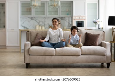 Happy calm Indian mom and little son kid sitting on couch, keeping lotus pose, zen hand gesture, meditating, practicing home yoga, home yoga, mental control, mindfulness, relaxing, exercising