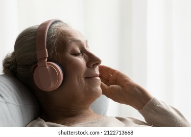 Happy Calm Relaxed Older Woman In Big Headphones Resting On Comfortable Couch, Listening To Music For Relaxation, Meditation, Enjoying Break, Leisure Time At Home, Smiling With Closed Eyes