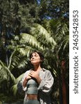 Happy calm young Hispanic woman holding hands on chest meditating doing yoga breathing exercises with eyes closed feeling gratitude, mental balance standing in green nature tropical park. Vertical.