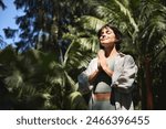 Happy calm young Hispanic woman holding hands in namaste meditating doing yoga breathing exercises with eyes closed feeling peace of mind, mental balance standing in green nature tropical park.