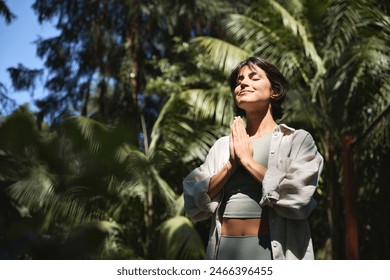 Happy calm young Hispanic woman holding hands in namaste meditating doing yoga breathing exercises with eyes closed feeling peace of mind, mental balance standing in green nature tropical park. - Powered by Shutterstock