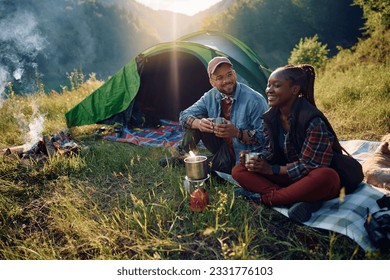 Happy camper and his African American girlfriend drinking tea while sitting on front of their tent in nature. - Powered by Shutterstock