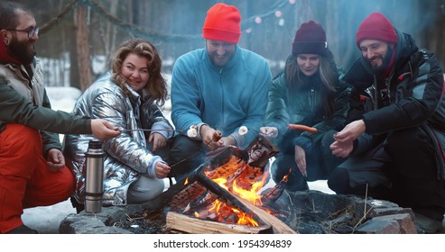 Happy Campers Sitting By Fire And Roasting Marshmallows And Sausages On Stick In Winter Forest. Young Friends Eating And Resting Near Campfire Hiking Together In Woods