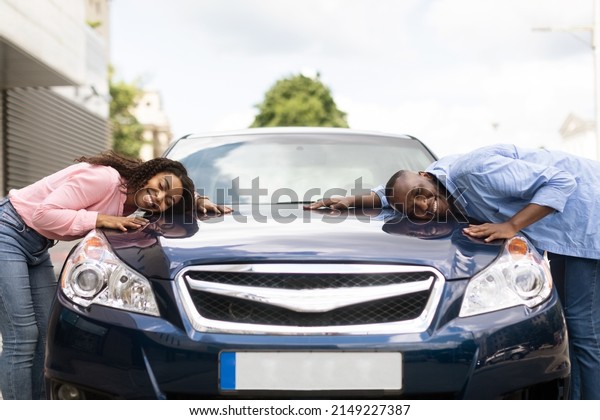 Happy Car Buyers, New Owners Concept. Portrait Of\
Excited Young African American Couple Touching Hugging New Luxury\
Automobile After Purchase In Dealership Showroom Standing Outdoors.\
Dreams Come True