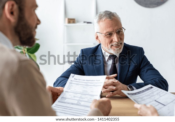 happy car dealer in glasses sitting with\
clenched hands near customers with contracts\
