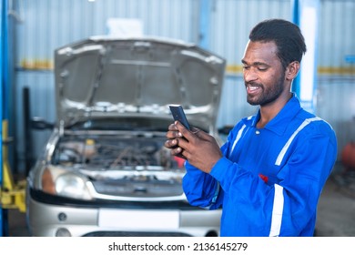 Happy Car Mechanic Busy Using Mobile Phone At Garage In Front Of Repair Car With Opened Hood - Concept Of Using Technology, Finding Customers Online And Relaxation.