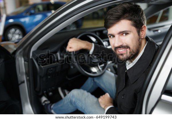 Happy car owner\
at dealership. Handsome young man sitting at front seat of the car\
looking at camera and\
smiling