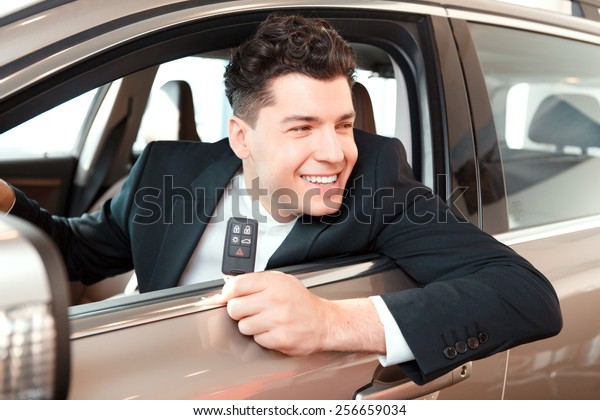 Happy car owner. Portrait of a smiling\
handsome man in suit sitting on drivers place in car and looking\
through the car window in car\
dealership
