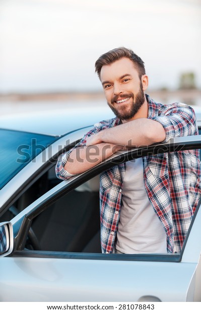 Happy car\
owner. Happy young man leaning at the door of his car and smiling\
at camera while standing\
outdoors