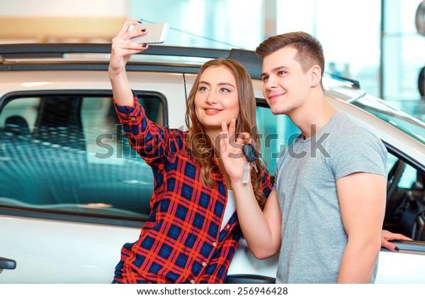 Happy car owners.\
Young couple standing near the car at the dealership and holding a\
key while making a selfie