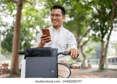 A happy, carefree Asian millennial businessman is checking messages on his smartphone while he is on a bike, riding a bike to work in the morning. modern city life and transportation concepts - Powered by Shutterstock