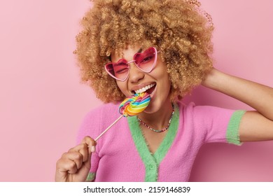 Happy carefree curly haired woman cannot live without sweets bites multicolred candy wears heart shaped sunglasses and t shirt isolated over pink background. Taste rainbow. Sweet life concept