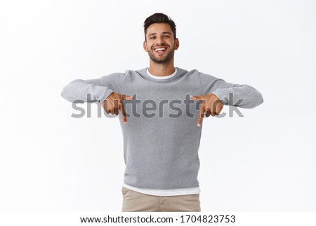 Happy carefree, friendly handsome man with bristle in grey casual sweater, joyfully talking, give advice, recommend promo as pointing bottom, indicate down link, standing white background