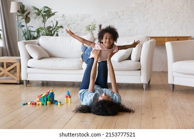 Happy carefree little Black kid playing airplane with mom, looking at camera, smiling. Mother lifting daughter child with flying hands in air, resting on heating floor, exercising with excited girl - Shutterstock ID 2153777521