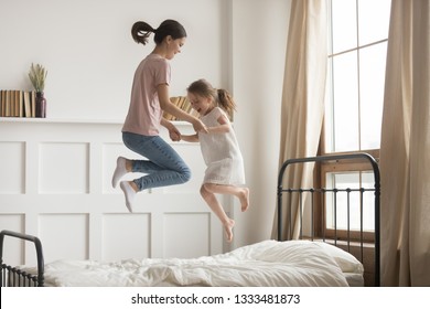 Happy carefree mom baby sitter and cute kid girl holding hands jumping on bed together, family mother playing having fun with little child daughter laughing feeling joy fly in air at home in bedroom