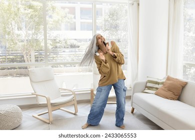 Happy carefree senior Latin woman singing at microphone object at home, dancing alone in living room, having fun, enjoying active leisure in cozy white colored home interior - Powered by Shutterstock