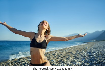 Happy Carefree Woman In Black Bikini Standing Against Blue Sky At Beach With Arms Outstretched, Feeling The Breeze And Freedom