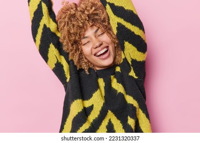 Happy carefree woman keeps eyes closed raises arms and dances carefree has upbeat mood expresses positive sincere emotions wears casual black and yellow jumper isolated over pink background. - Shutterstock ID 2231320337