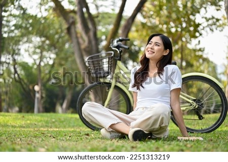 Happy and carefree young Asian woman in casual clothes sits on the grass, looking at the beautiful greenery of nature view, taking a rest after cycling in the park.