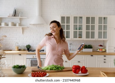 Happy carefree young attractive woman holding cellphone in hands, dancing to disco music from mobile player application, having fun preparing healthy fresh vegetarian food alone in modern kitchen.