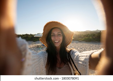 A happy carefree young female tourist with a straw hat is making selfie or technology video call with a phone to a friends or parents on a desert offshore seaside in a sunny day during vacation trip.