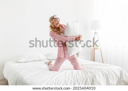 Happy carefree young woman with smile on face has fun on bed with white bedding in cozy bedroom, attractive lady in pajama enjoying morning on weekend at home. Female in pajamas hugging pillow