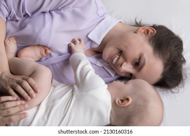 Happy Caring Devoted New Mom Enjoying Relaxation With Infant Kid, Hugging, Cuddling, Caressing Baby, Smiling, Talking To Child, Resting On Bed With Pale Sheet. Motherhood Concept