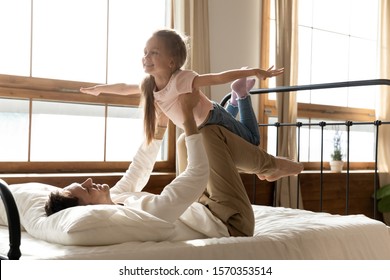 Happy caring father holding lifting cute little preschool child daughter up playing plane game on bed at home, funny kid girl flying bonding having fun with dad enjoying morning lifestyle in bedroom - Powered by Shutterstock