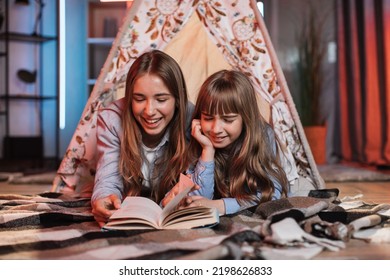 Happy Caring Nanny Or Older Sister Reading Fairytale To Her Younger Sister During Evening Time Outside Teepee Tent Lying On The Floor At Dark Cozy Room.