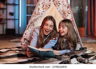 Happy Caring Nanny Or Older Sister Reading Fairytale To Her Younger Sister During Evening Time Outside Teepee Tent Lying On The Floor At Dark Cozy Room.