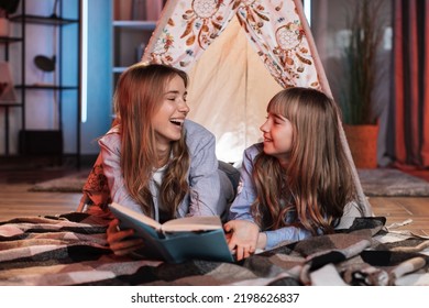 Happy Caring Nanny Or Older Sister Reading Fairytale To Her Younger Sister During Evening Time Outside Teepee Tent Lying On The Floor At Dark Cozy Room.