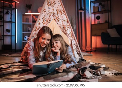 Happy Caring Nanny Or Older Sister Reading Fairytale To Her Younger Sister During Evening Time Outside Teepee Tent Lying On The Floor At Dark Cozy Room.