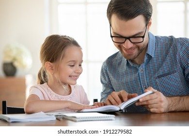 Happy caring young dad and little daughter sit at table at home learn read book prepare for school together. Smiling loving Caucasian father and small girl child study do homework. Education concept.