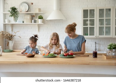 Happy Caring Young Mother Sit At Table In Modern Kitchen Cooking Together With Little Preschooler Daughters, Millennial Caucasian Mom Or Nanny Prepare Meal Dinner Salad With Small Children At Home