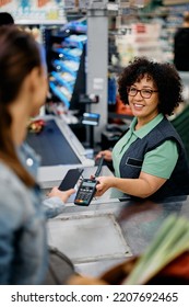 Happy Cashier Holding Credit Card Reader While Customer Is Paying With Smart Phone At Supermarket. 