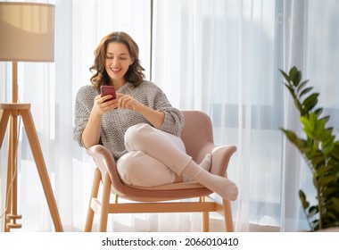 Happy casual beautiful woman is using a phone sitting on armchair at home. - Powered by Shutterstock