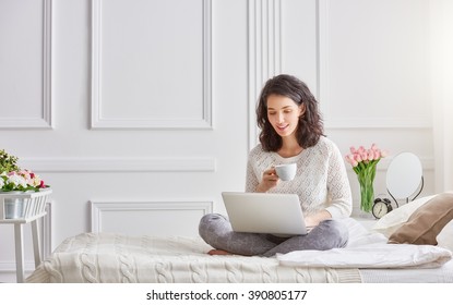 Happy casual beautiful woman working on a laptop sitting on the bed in the house.