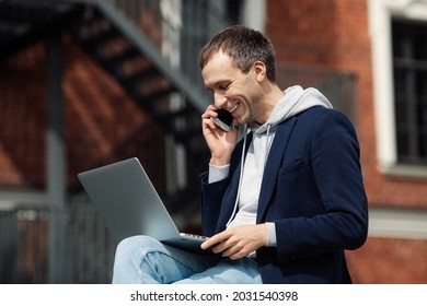 Happy Casual Entrepreneur Talk On Smartphone With Colleague Holding Laptop Analyze Growth In Marketing Sales Report On Mobile Phone Conversation Outside Office. Freelance Worker Manager Work Remotely