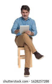 Happy Casual Man Reading And Writing On His Tablet While Sitting On A Chair On White Studio Background