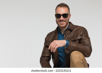 Happy Casual Man Is Resting His Elbow On His Knee And Wearing A Brown Leather Jacket And Sunglasses On Gray Studio Background