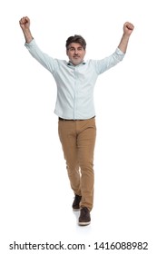 Happy Casual Old Man Stepping Forward And Celebrating With Both Fists In The Air While Wearing A White Shirt On White Studio Background