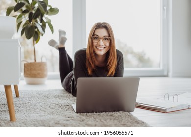 Happy Casual Young Woman Lying On The Floor At Home With Her Laptop Looking Over The Top At The Camera With A Friendly Smile In A Low Angle High Key View