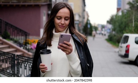 Happy Caucasian Business Woman In White Sweater And Black Leather Jacket Walking Down The Street To Her Company Office Holding Coffee And Smartphone.