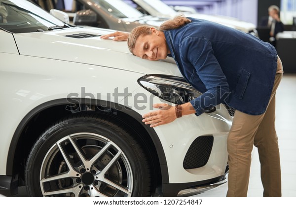 Happy caucasian businessman hugging bonnet of\
his new white car in dealership, being completely satisfied with\
expensive purchase