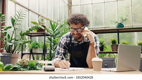 Happy Caucasian businessman talking on cellphone while standing in apron in small floral center and writing down order details. Joyful male florist calling on smartphone at work. Own business concept - Shutterstock ID 1852040428