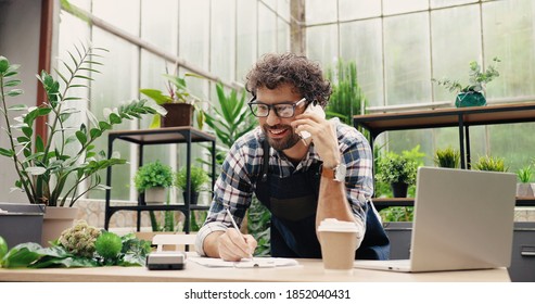 Happy Caucasian businessman talking on cellphone while standing in apron in small floral center and writing down order details. Joyful male florist calling on smartphone at work. Own business concept