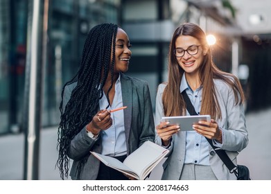 Happy Caucasian Businesswoman Using Tablet While Walking With Her African American Female Colleague. Multiracial Business People Outside Talking About Their Work Progress. Diversity. Copy Space.