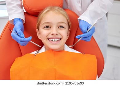 Happy Caucasian Child Girl Came To See The Dentist. Kid Sits In Dental Chair. Dentist In Blue Gloves Bent Over Her, Top View. Happy Patient And Dentist Concept. Adorable Child Look At Camers Smiling